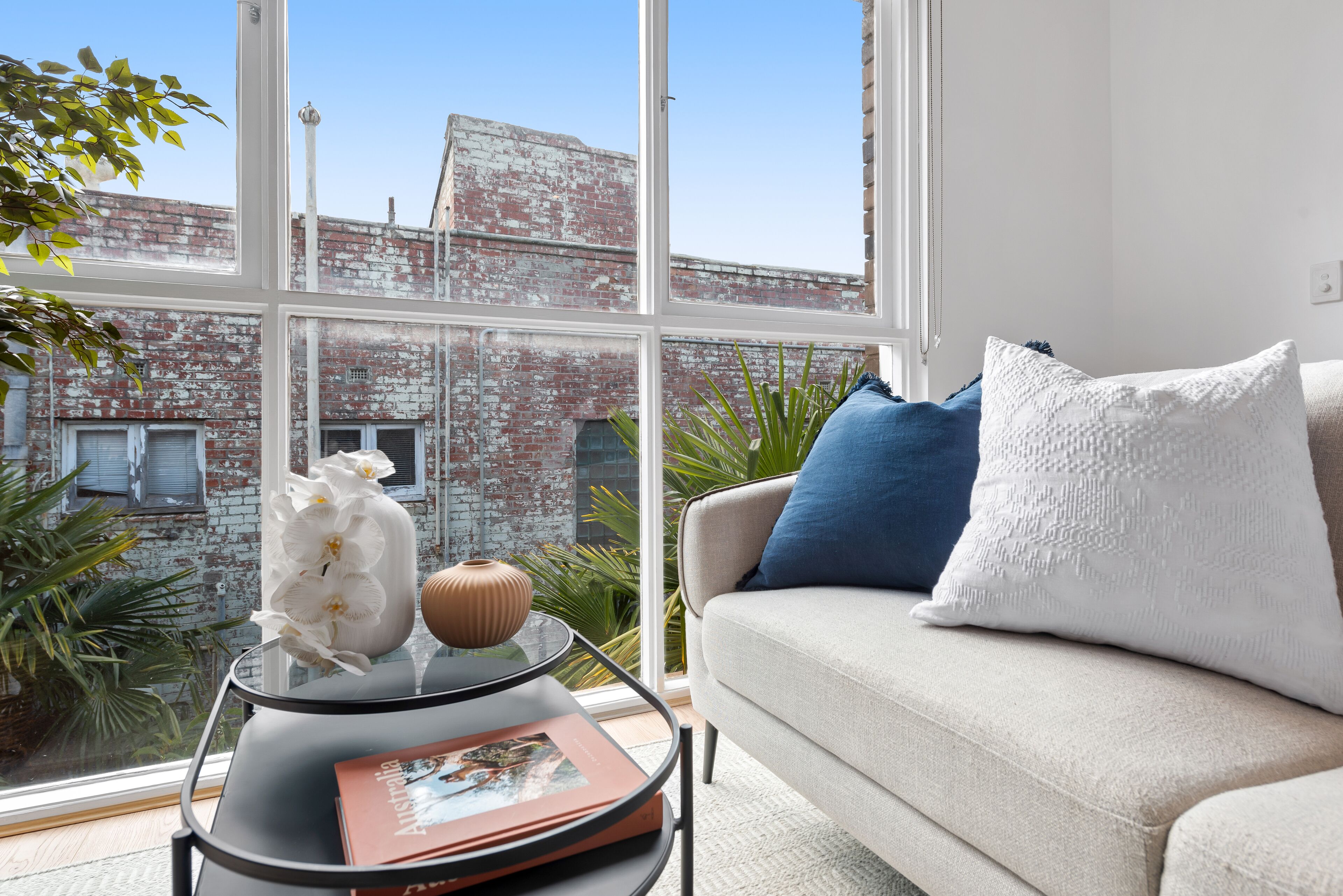 A modern living room with a large window, a sofa with blue and white cushions, and a glass coffee table with a book and decorative items.