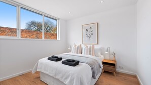Un dormitorio moderno con un ventanal grande, una cama con ropa de cama blanca, una mesita de noche con una lámpara e impresión botánica enmarcada en la pared.