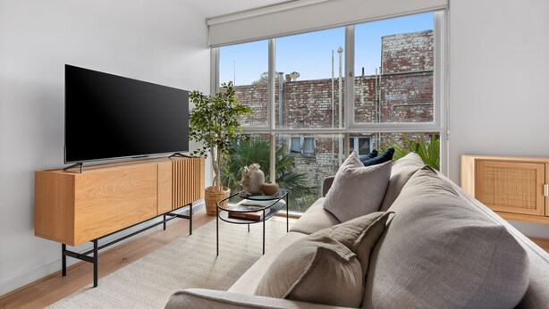 Un salón moderno con un televisor de pantalla plana grande, un mueble de madera, una mesa de centro de vidrio, un sofá color beige y vistas a un edificio de ladrillo a través de amplias ventanas.