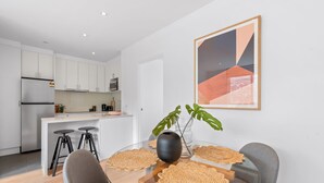 Una cocina moderna con armarios blancos, un refrigerador de acero inoxidable y un microondas. Hay un comedor con una mesa de vidrio, dos sillas y un florero con una planta.