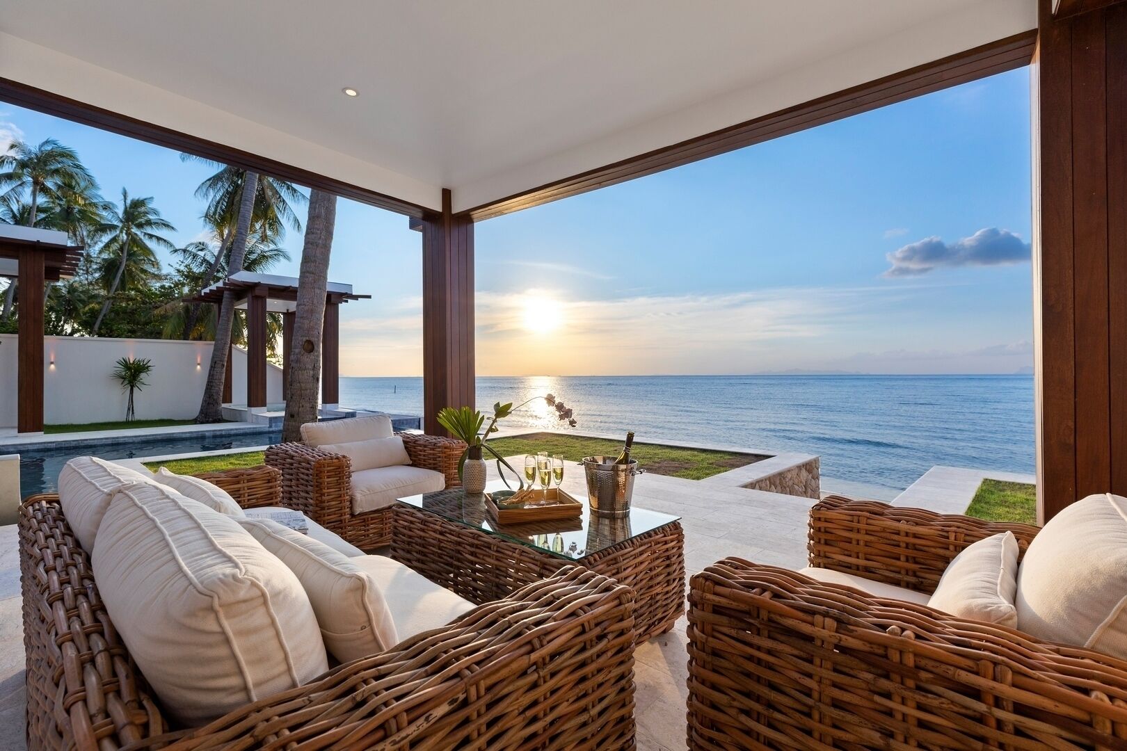 Un espace extérieur moderne, aménagé avec des meubles en osier et une table en verre, offrant une vue sur une piscine et l’océan.