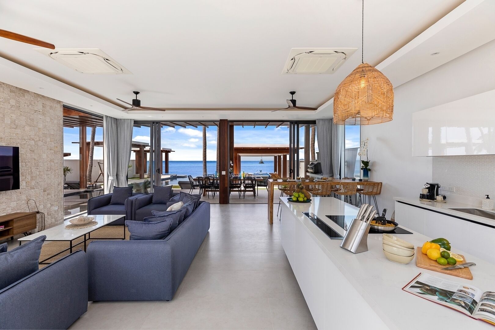 Un salon moderne avec un grand canapé, un coin repas offrant une vue sur la mer et une cuisine dotée d’un grand îlot central.