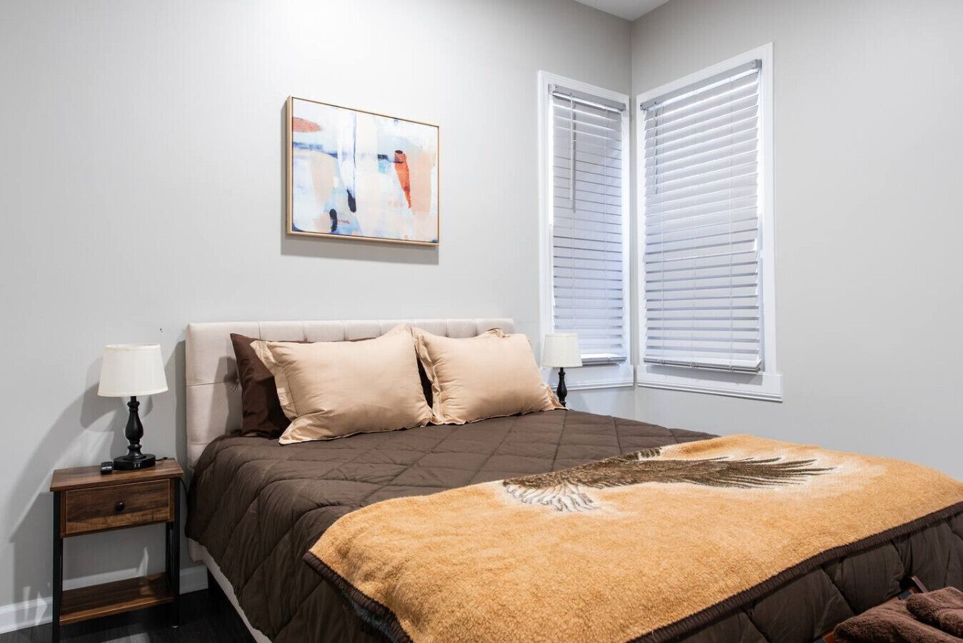A bedroom with a bed, two bedside tables, a nightstand, a painting, and two windows with blinds.