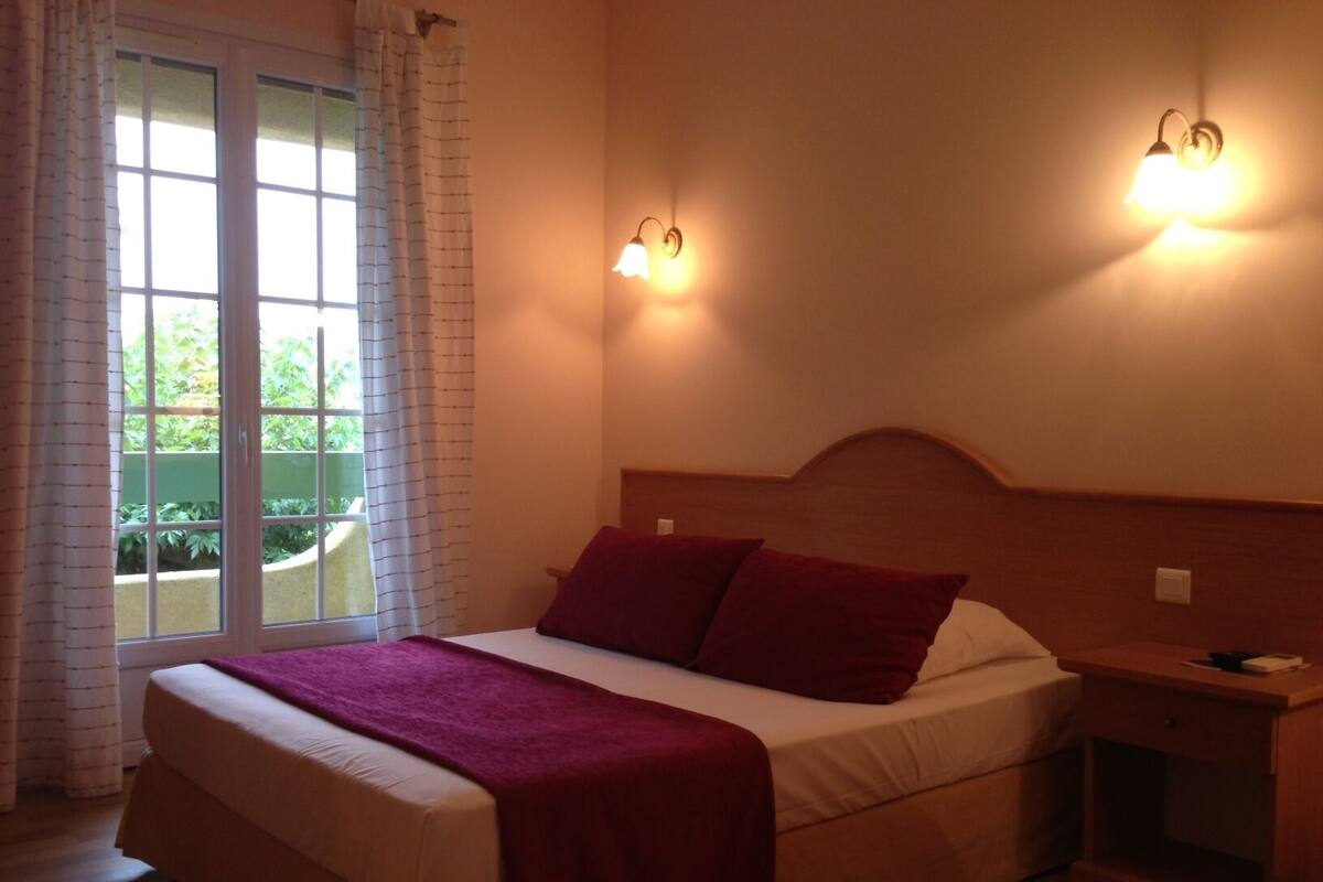 Un dormitorio con cama, mesita de noche y ventana con cortinas.