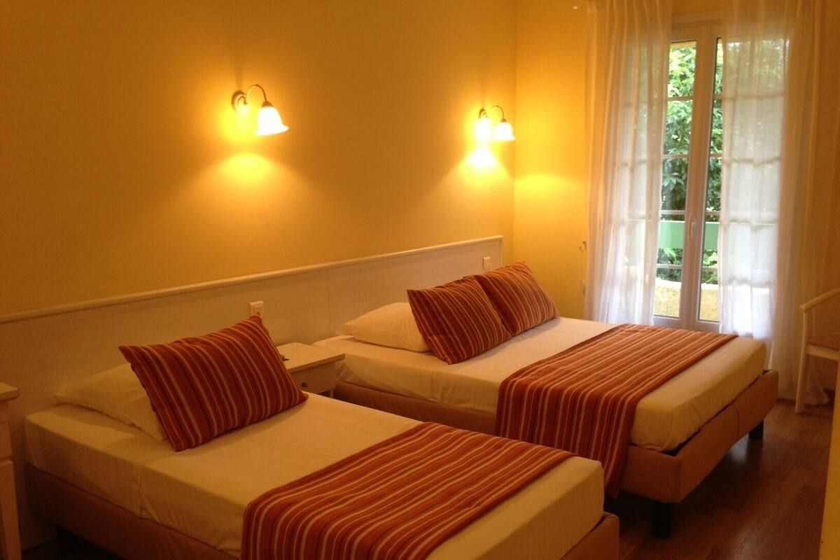 Habitación de hotel con dos camas individuales, cada una con almohada y manta a rayas, cabeceros blancos y dos lámparas fijadas a la pared.