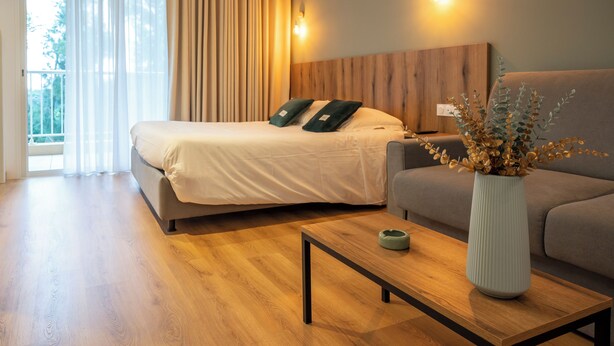 Una habitación de hotel moderna con una cama, un sofá, una mesa de centro de madera y un florero con flores secas.