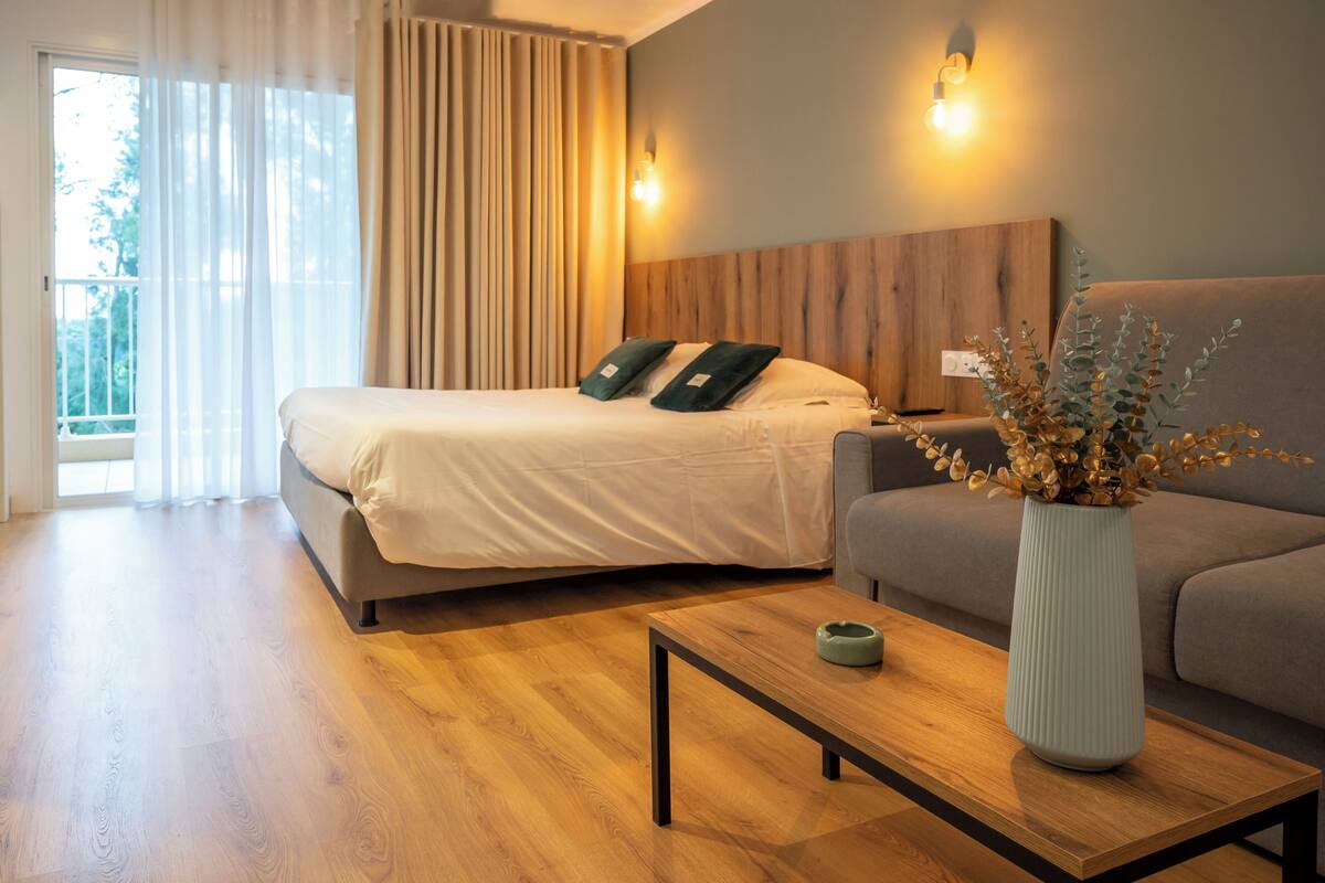 Una habitación de hotel moderna con una cama, un sofá, una mesa de centro de madera y un florero con flores secas.