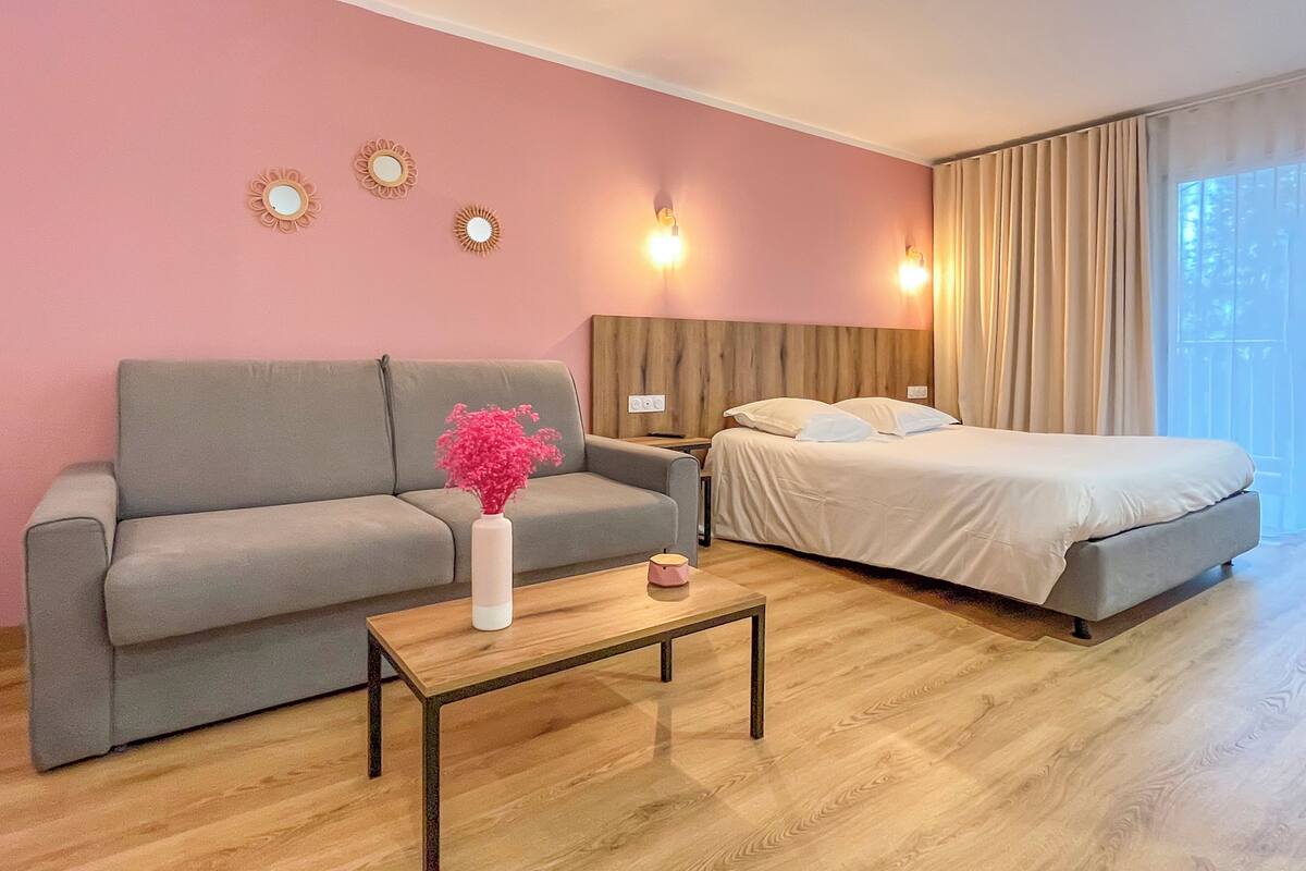 Una habitación con un sofá gris, una mesa de centro de madera, una cama con ropa de cama blanca y una pared rosa con espejos decorativos.