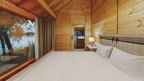 A wooden cabin room with a bed, a nightstand, and a window overlooking a body of water. - Zaga Zaga Sat (Cosmesti)
