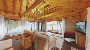 A cabin interior with a sofa, dining table, and TV. Large windows offer views of trees and water. - Zaga Zaga Sat (Cosmesti)