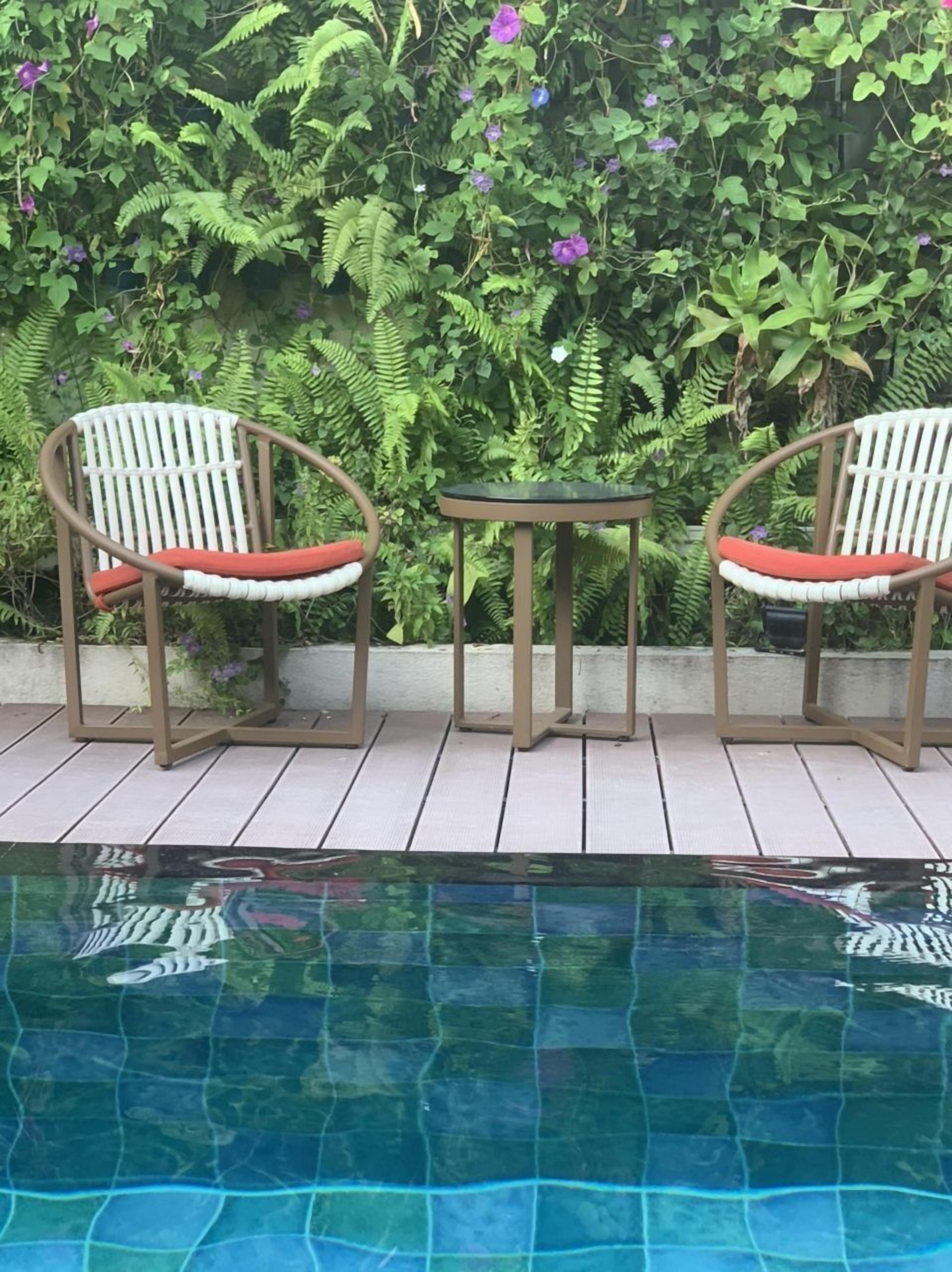 A poolside area with two chairs and a small table, surrounded by lush greenery and a pool.