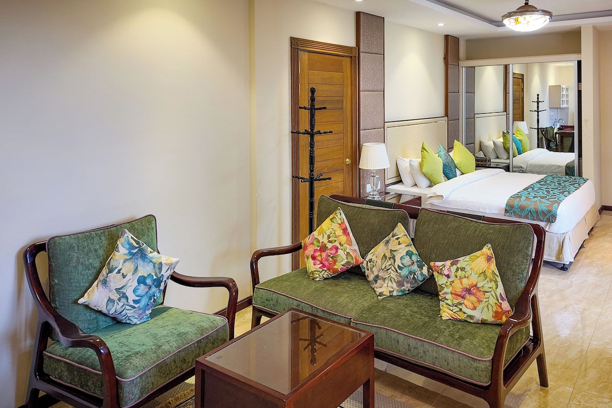 A hotel room with a green sofa set, a wooden coffee table, and a bed with floral bedding.
