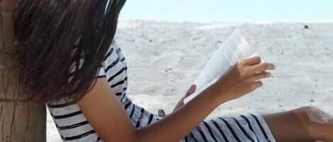 A person sitting on a beach, reading a book, with a clear view of the ocean and a palm tree in the background.