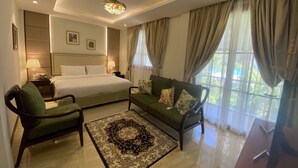 Habitación de hotel con cama, sofá, dos sillones, alfombra decorativa, mesita de noche con lámpara e iluminación en el techo.