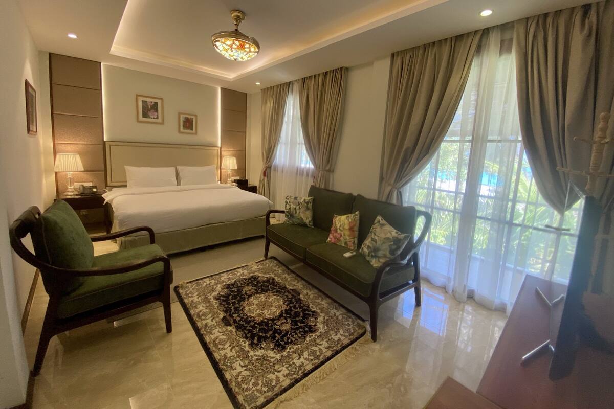 A hotel room with a bed, a sofa, two armchairs, a decorative rug, a nightstand with a lamp, and a ceiling light fixture.