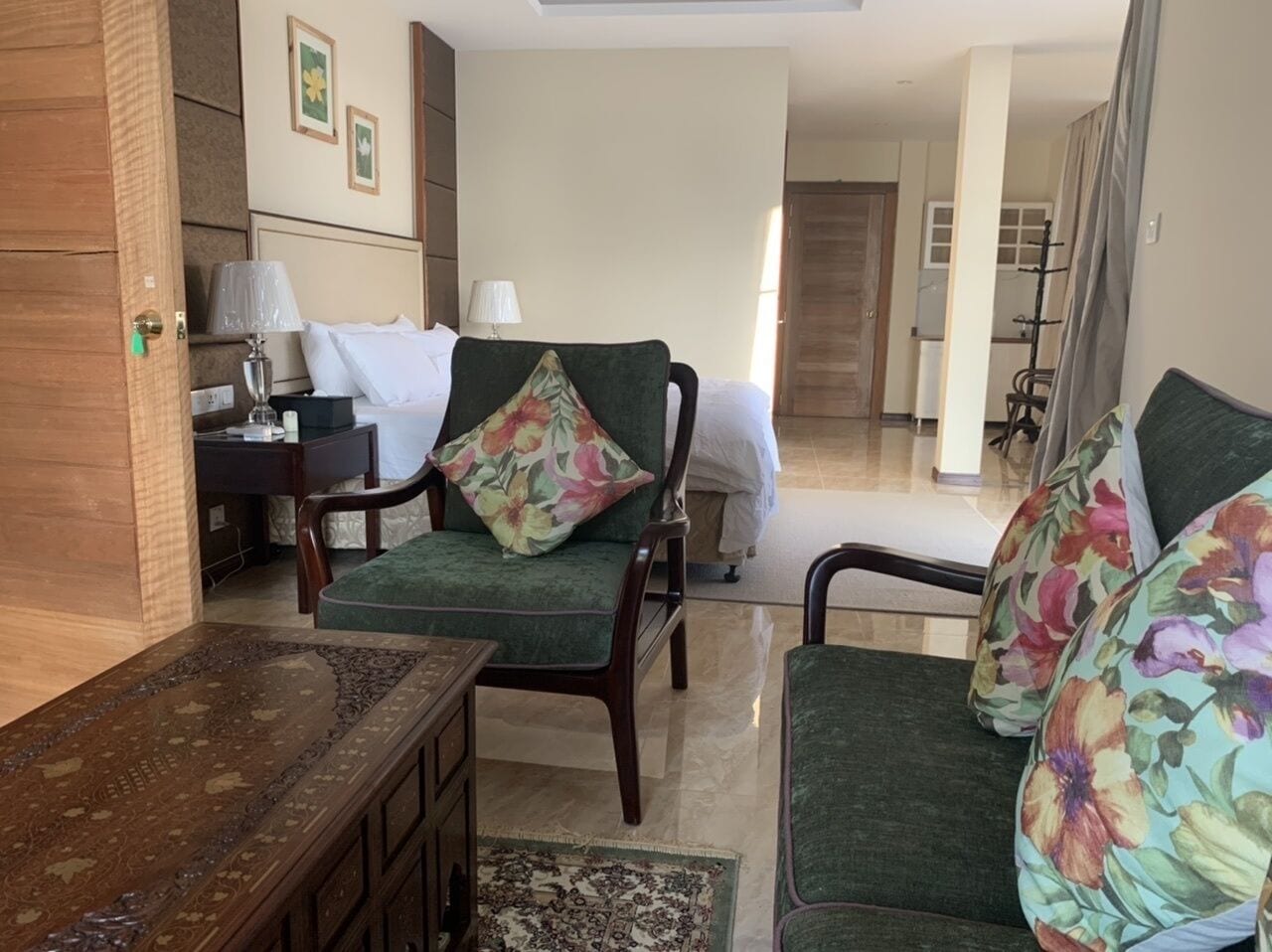 A hotel room with a bed, two green armchairs with floral cushions, a wooden side table, and a lamp.