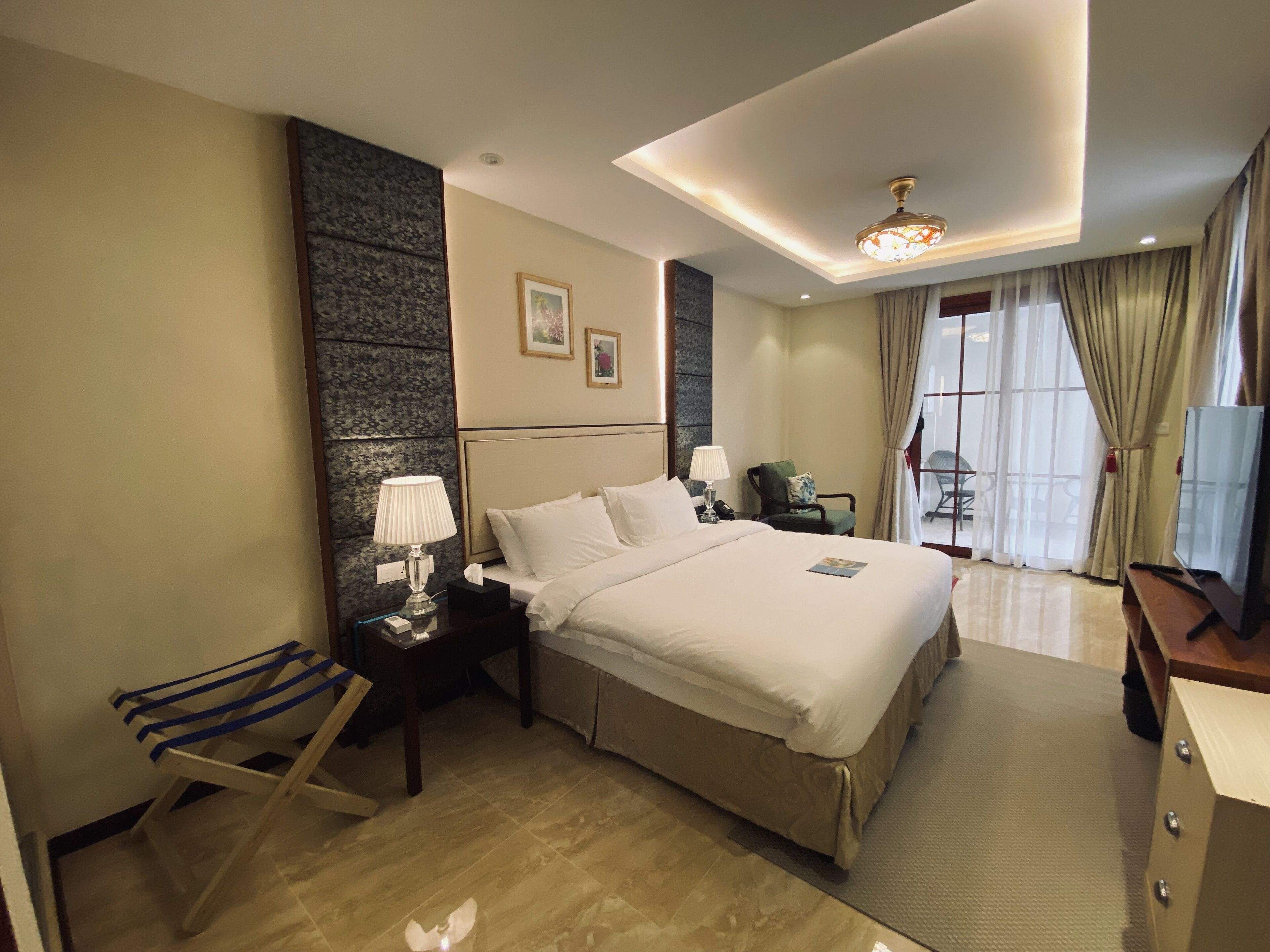 Habitación de hotel con una cama grande, un escritorio con lámpara, una silla, televisión y ventana con cortinas.