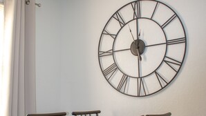 Een eethoek met een tafel en vier stoelen, een hangklok aan de muur, en een raam met gordijnen.