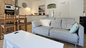 A modern living room with a grey sofa, wooden chairs, a white table, and a kitchen area with a microwave and stove. - Haut du village, T2 + alcôve 5p. (Cauterets)