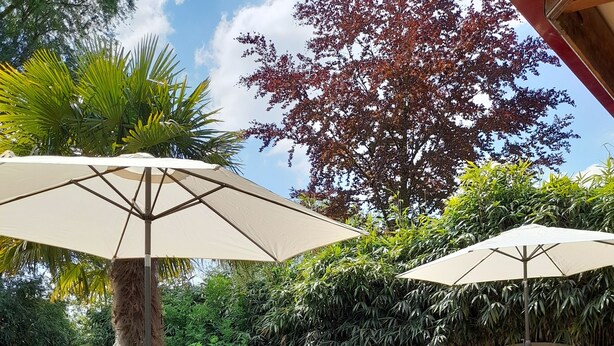 Un patio al aire libre con terraza de madera, una mesa con cubierta de vidrio, sillas de mimbre y dos grandes sombrillas.