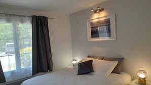 Un dormitorio con cama, una mesita de noche con lámpara, un cuadro enmarcado en la pared y una ventana con cortinas.