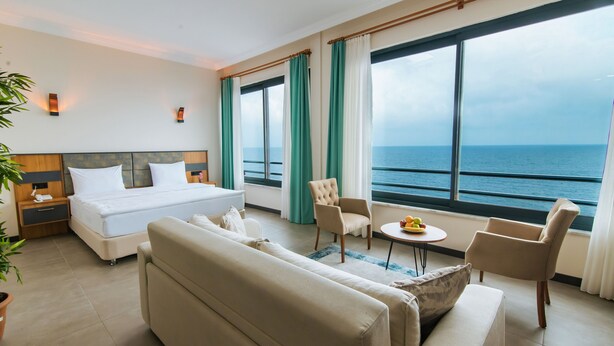 A hotel room with a bed, sofa, armchair, and a table with fruit. There is a large window with a view of the sea. - Fevzi Hoca Hotel (Akçaabat)
