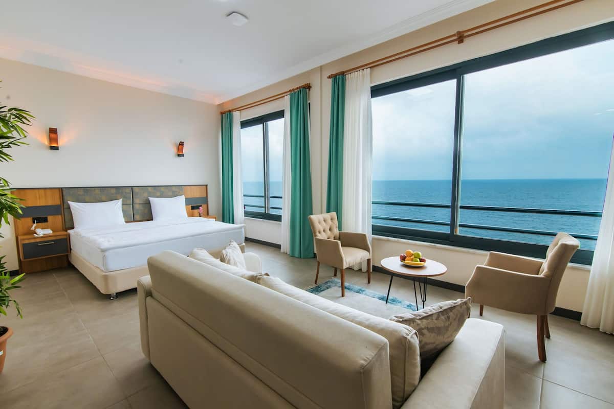 Quarto de hotel com cama, sofá, poltrona e mesa com frutas. Há uma janela grande com vista para o mar.