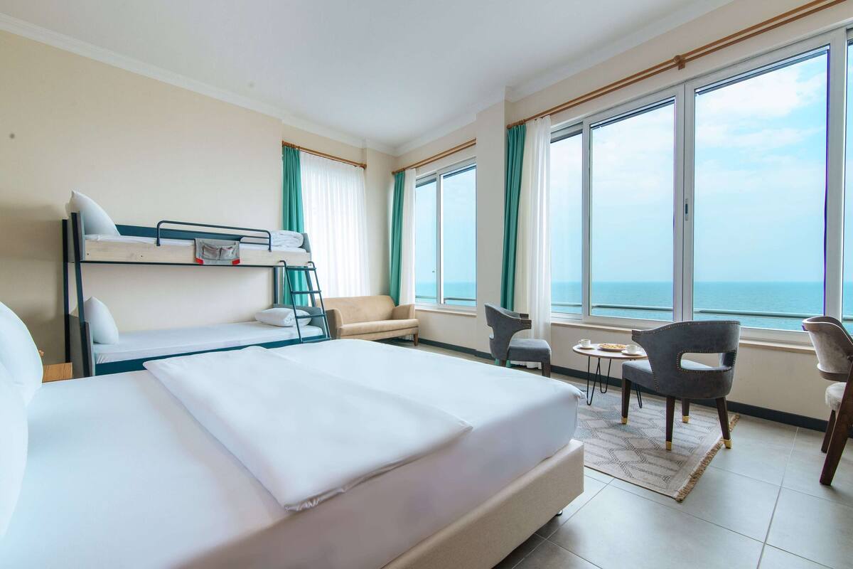 Quarto de hotel com uma cama grande, um sofá, uma mesa pequena e uma cadeira. Há grandes janelas com cortinas, e vista para o mar.
