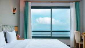 A hotel room with a large bed, a desk with a chair, and a view of the sea through a large window. - Fevzi Hoca Hotel (Akçaabat)