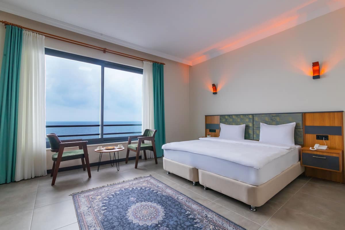 Quarto de hotel com uma cama grande, uma escrivaninha com cadeira, uma mesa pequena e vista para o mar através de uma janela ampla.