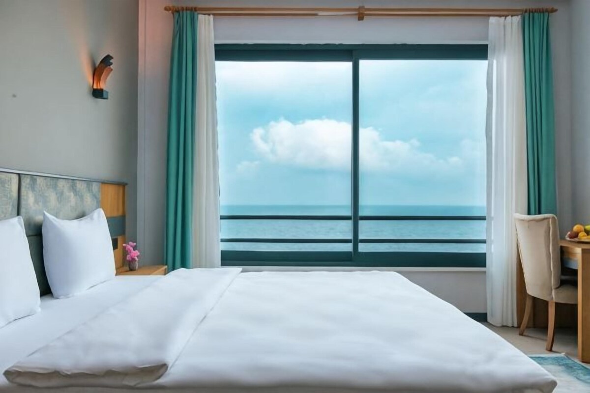 A hotel room with a large bed, a view of the sea, a desk with a chair, and a nightstand with a lamp.