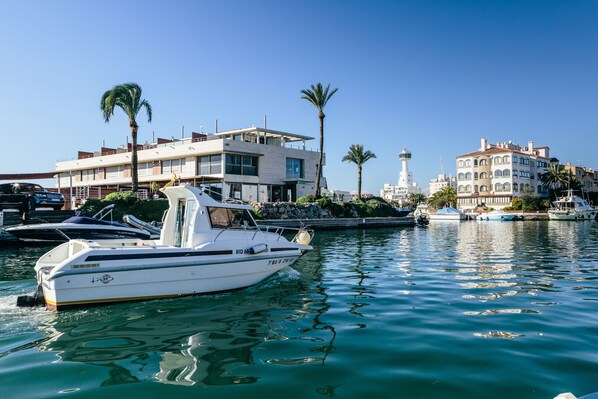 Un puerto deportivo con barcos amarrados, un edificio moderno con balcones y un faro al fondo.