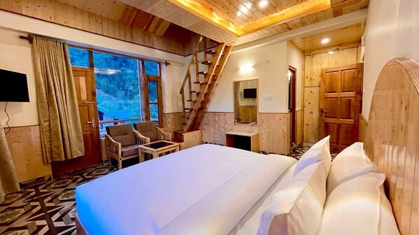 A wooden cabin-style room with a large bed, a desk, and a chair. There is a staircase leading to an upper level, and a window with curtains.