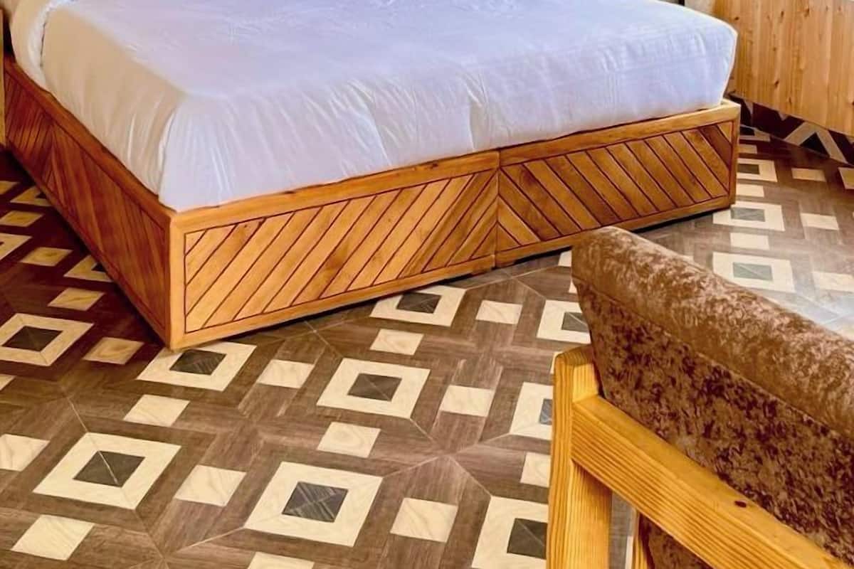 Un lit en bois avec une literie blanche, dans une pièce dont le sol est recouvert d’un motif décoratif et qui possède une fenêtre avec des rideaux.