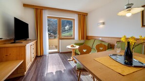 A room with a wooden desk, a television, a dining table with a vase and glasses, a sofa, and a window with curtains. - Apartment 'Obermoarhof Hönigspitze OG Deluxe' with Mountain View, Shared Garden and Wi-Fi (Lagundo)