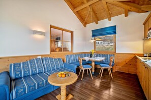 A room with a wooden ceiling, a blue sofa with patterned cushions, a dining table with chairs, and a small round table with fruit.