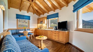 Quarto aconchegante com sofá, mesa de jantar, TV, armários de madeira e vista para as montanhas.