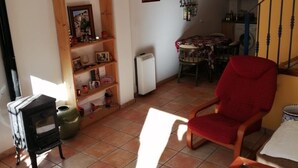 Un salon avec une cheminée, un fauteuil rouge, une table à manger recouverte d’une nappe à motifs, et un coin cuisine à l’arrière-plan.