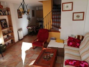 Un salon avec un fauteuil rouge, un canapé, une table basse, un coin repas avec une table et des chaises, un escalier et une cuisine à l’arrière-plan.