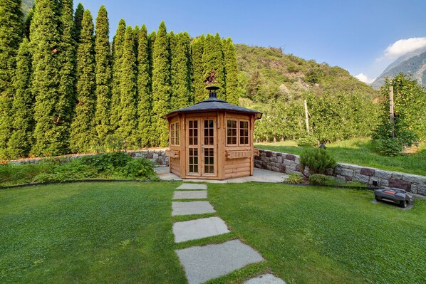 A wooden gazebo with a stone pathway leading to it, surrounded by lush greenery and a mountainous backdrop.