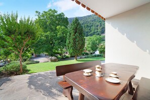 Une terrasse avec une table dressée pour deux personnes, donnant sur un jardin arboré et surmontée d’une vue sur une montagne.