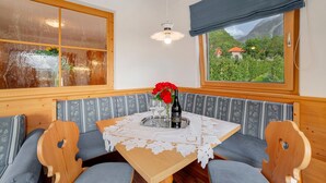 A wooden dining table with a lace tablecloth, a bottle of wine, and a vase with red flowers. There are two upholstered chairs with a floral pattern. - Apartment 'Obermoarhof Mutspitze' with Mountain View, Shared Garden and Wi-Fi (Lagundo)