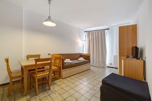 Una habitación con un sofá de madera, una mesa de comedor de madera con sillas, un pequeño otoman negro, un armario de madera y una lámpara en el techo.
