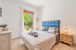A bedroom with a large bed, a nightstand, a wooden dresser, and a window with a view of greenery.