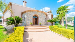Um edifício de pedra com porta de madeira, rodeado por vegetação bem cuidada e um caminho pavimentado.