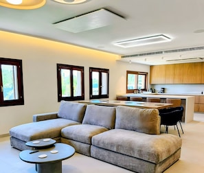 A modern living room with a grey sectional sofa, a small round coffee table, and a kitchen area with wooden cabinets and a central island. - Villa Levante Freshly Renovated for 2026 - With Wi-Fi, Air Conditioning, and Heated Pool (Sa Pobla)