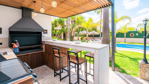 Uma cozinha externa com churrasqueira, banquetas e uma piscina ao fundo.