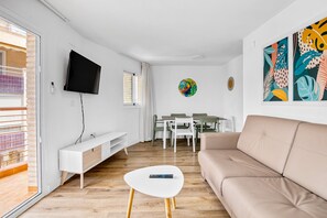 A modern living room with a sofa, a TV mounted on the wall, a dining area with chairs and a table, and a colorful wall decoration.
