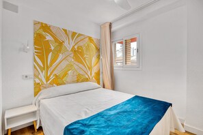 A bedroom with a bed, a bedside table, a window with curtains, and a wall with a leaf-patterned artwork.