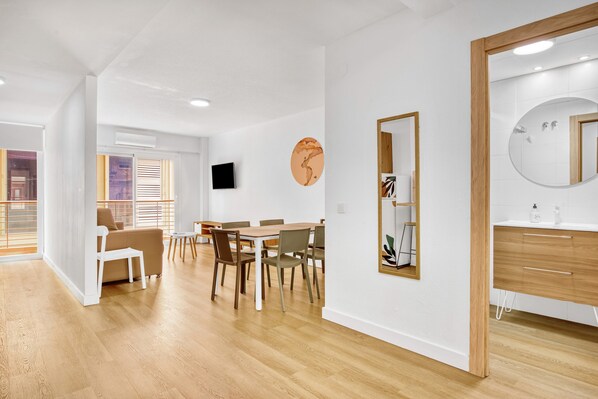 A modern hotel room with a wooden floor, a dining area with a table and chairs, a large mirror, and a TV on the wall.